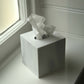 A square white marble tissue box cover with a white tissue paper partially pulled out, sitting on a windowsill next to a window.