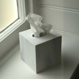 A square white marble tissue box cover with a white tissue paper partially pulled out, sitting on a windowsill next to a window.
