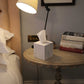 A white marble-patterned cube tissue box holder sitting on a round wooden bedside table next to a bed, with a lit lamp and books in the background.