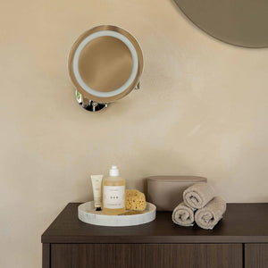A wall-mounted round vanity mirror with an integrated LED ring and chrome base, shown above a bathroom counter with rolled towels and toiletries in a warm beige room.