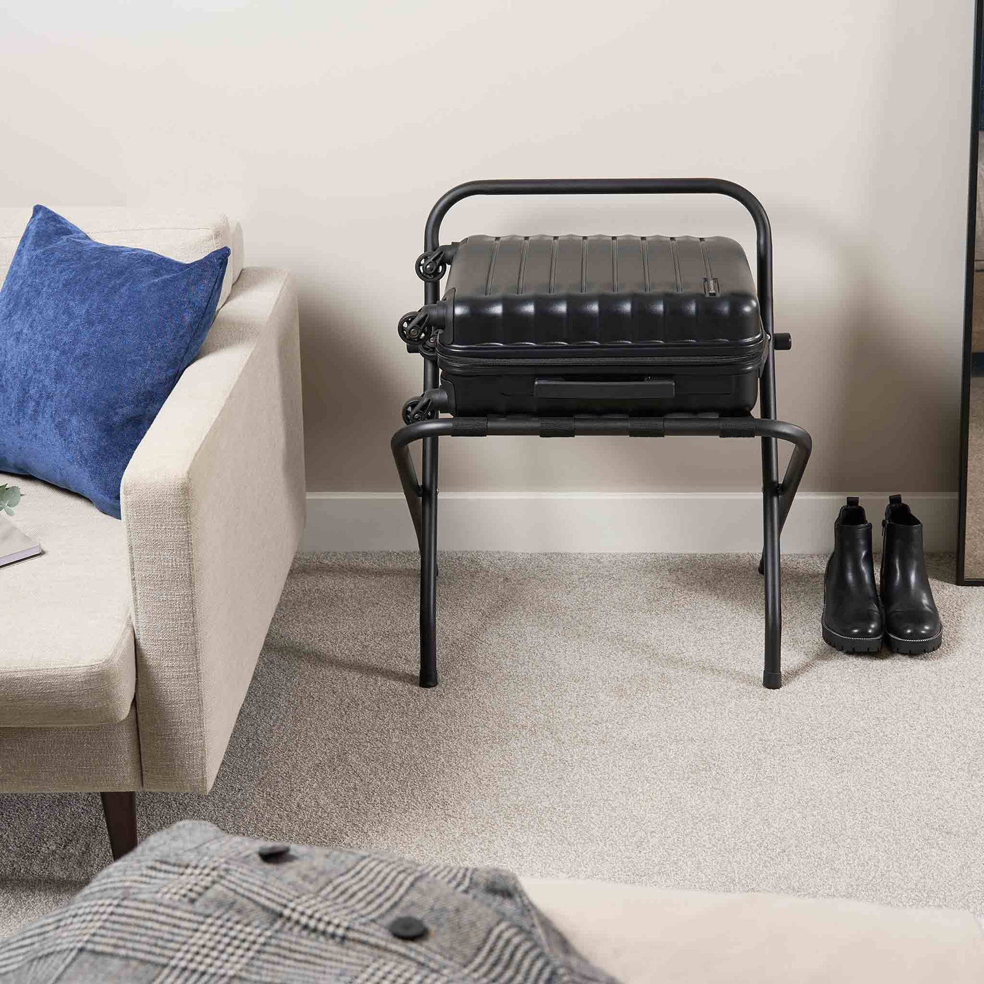 Corby Ashton matte black folding luggage rack with backrest holding suitcase in hotel guest room next to sofa and boots