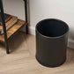 Black trash can on a wooden floor next to a bookshelf.