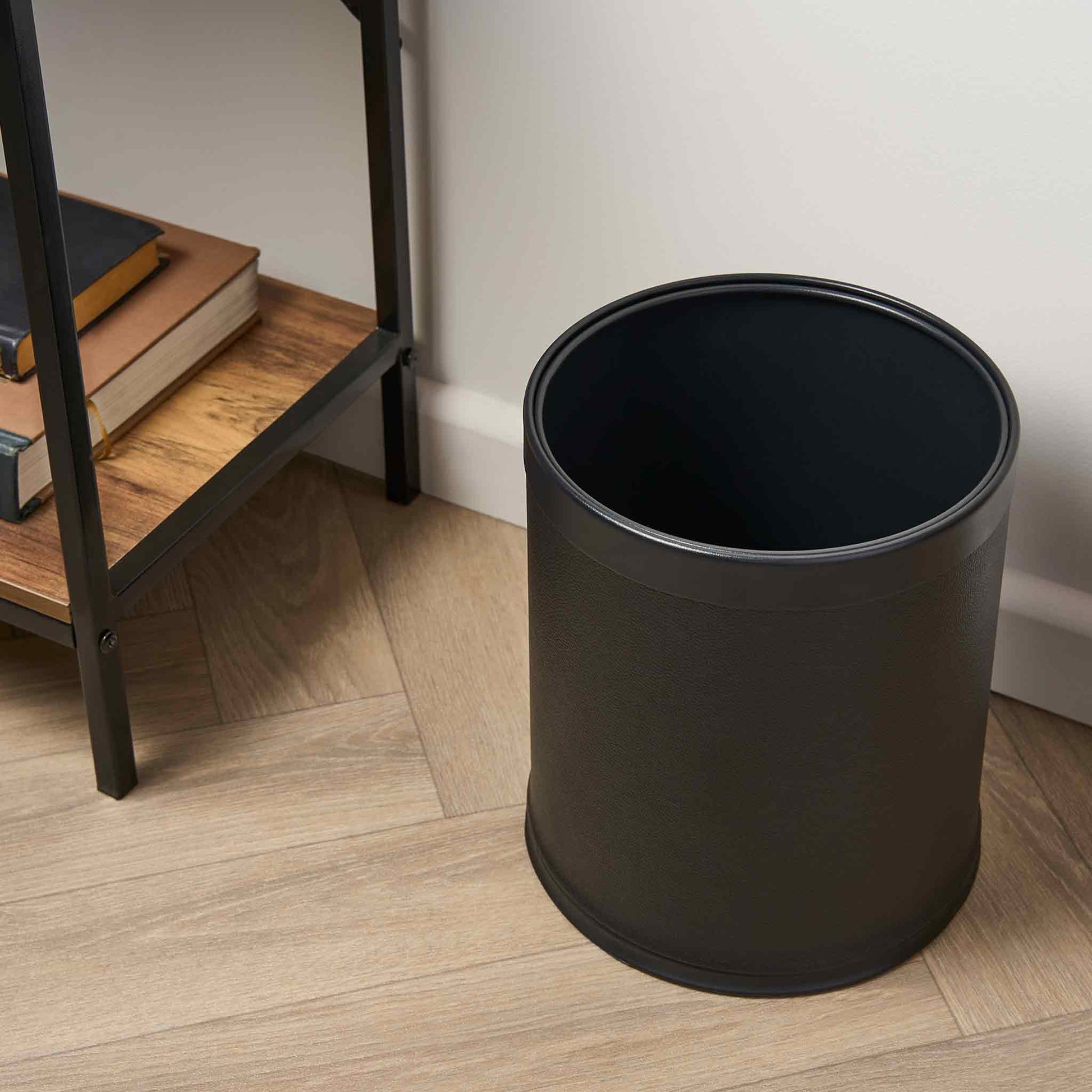 Black trash can on a wooden floor next to a bookshelf.