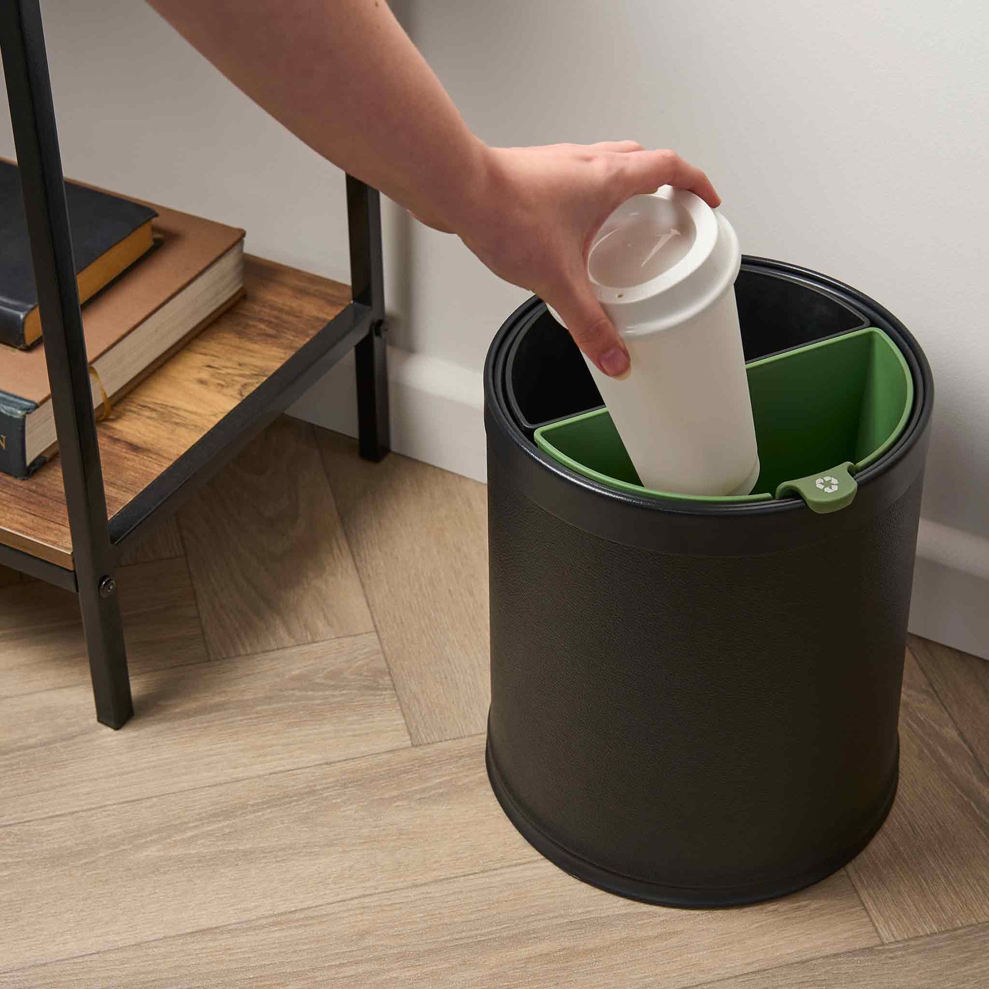 Person disposing of a coffee cup into a black and green trash can on a wooden floor.