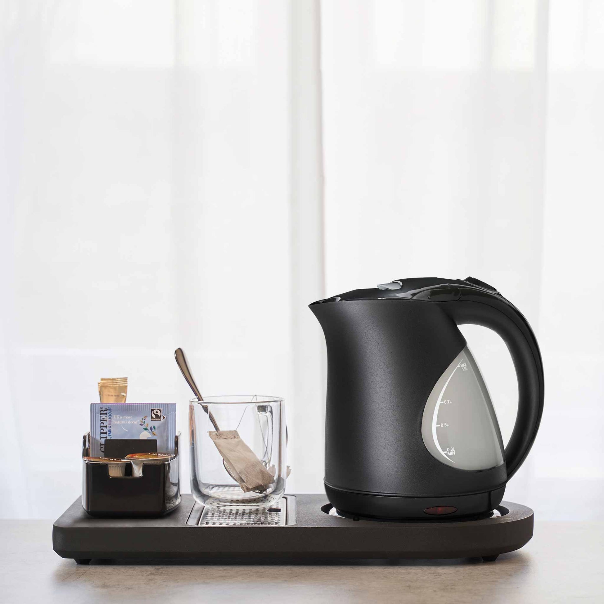 Corby Lancaster 1L black hotel kettle on hospitality tray with glass mug, tea sachets, and spoon, set against a light, minimalist background