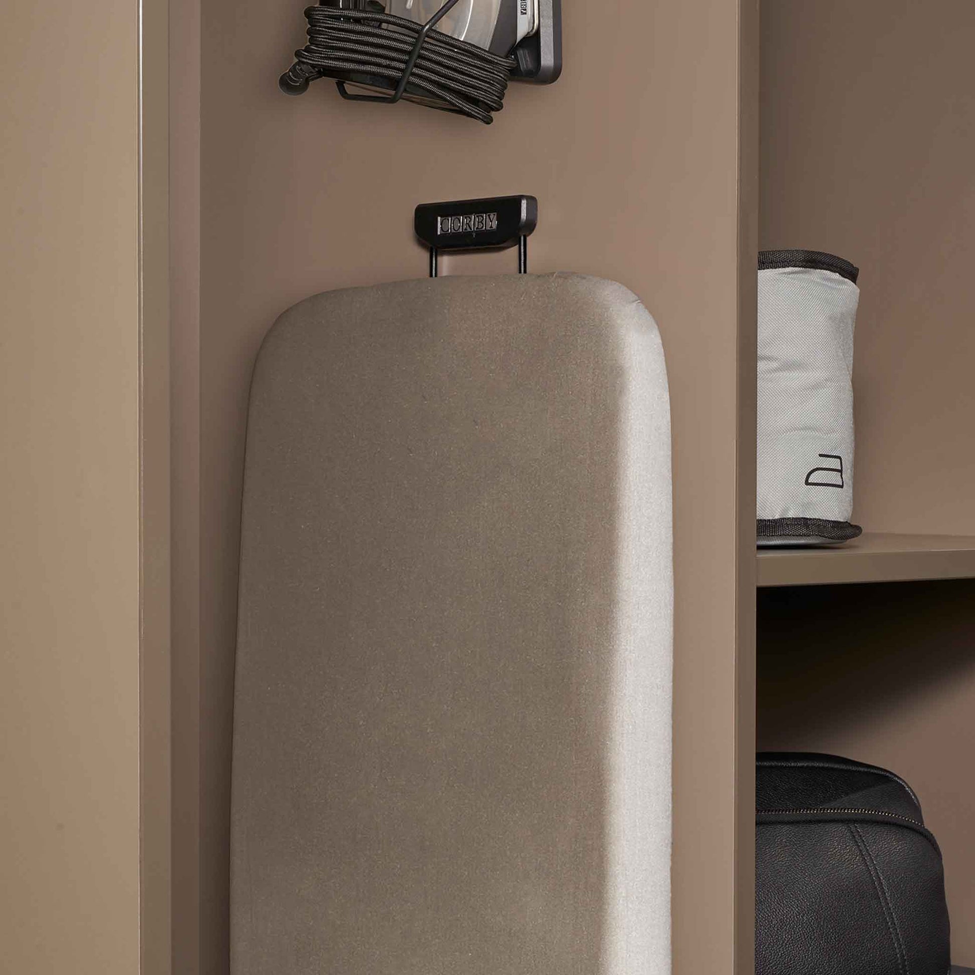 Corby Sherwood ironing board with light grey cover stored vertically in hotel wardrobe using compact black holder, iron mounted above