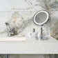 Corby Winchester LED vanity mirror with chrome stand on hotel bathroom counter beside toiletries, towel, and sink with chrome tap