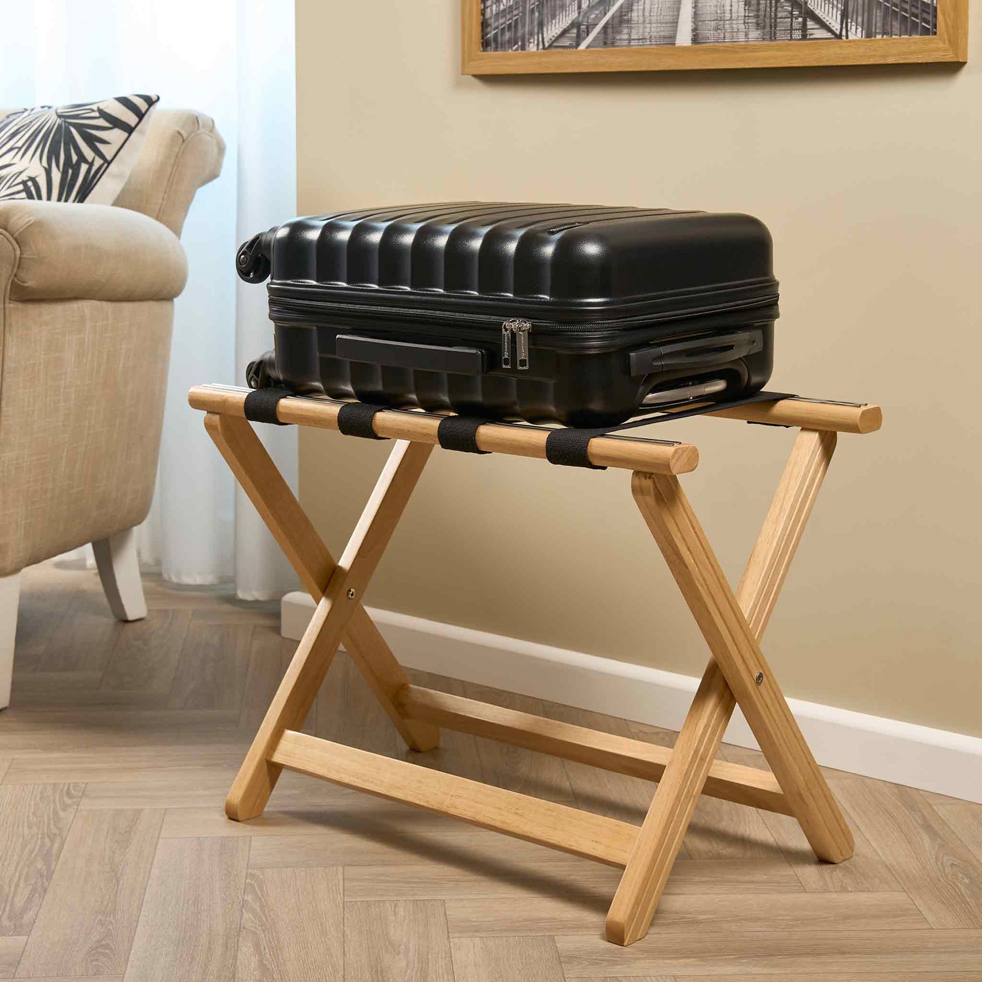 Corby York light wood hotel luggage rack with black fabric straps, holding hard-shell suitcase in guest room setting with neutral décor