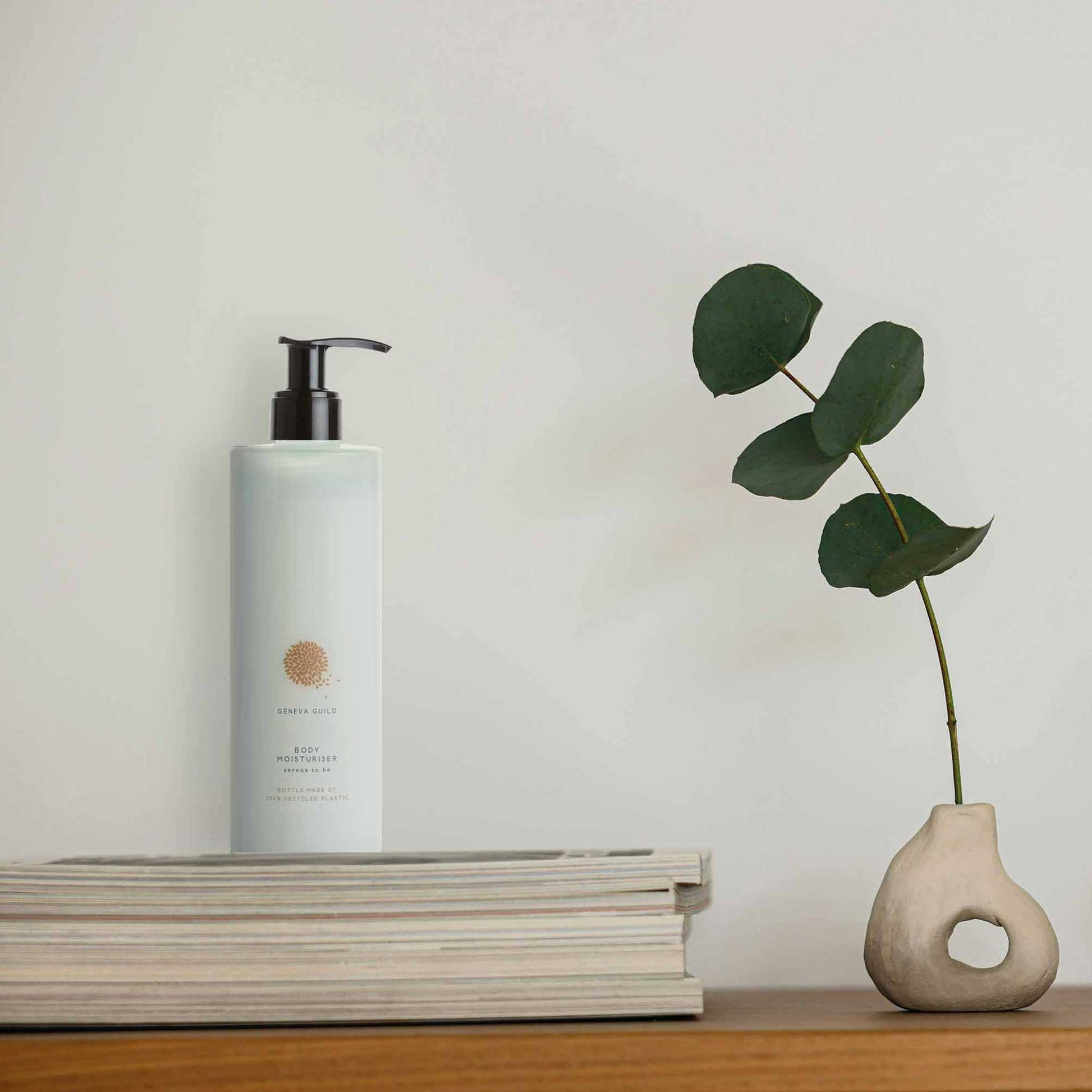 Geneva Guild body moisturiser 380ml pump dispenser placed on books beside sculptural vase and eucalyptus stem in a minimalist hotel or spa setting