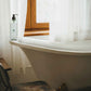 Geneva Guild body wash 380ml pump bottle placed beside freestanding bathtub in a rustic-style guest bathroom with natural light and wood-framed window