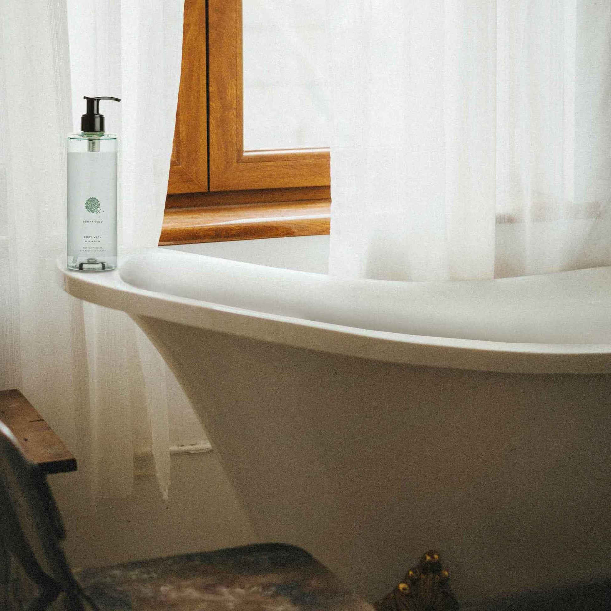 Geneva Guild body wash 380ml pump bottle placed beside freestanding bathtub in a rustic-style guest bathroom with natural light and wood-framed window