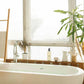Geneva Guild shampoo 380ml pump dispenser placed on windowsill beside freestanding bathtub in bright, plant-filled hotel bathroom