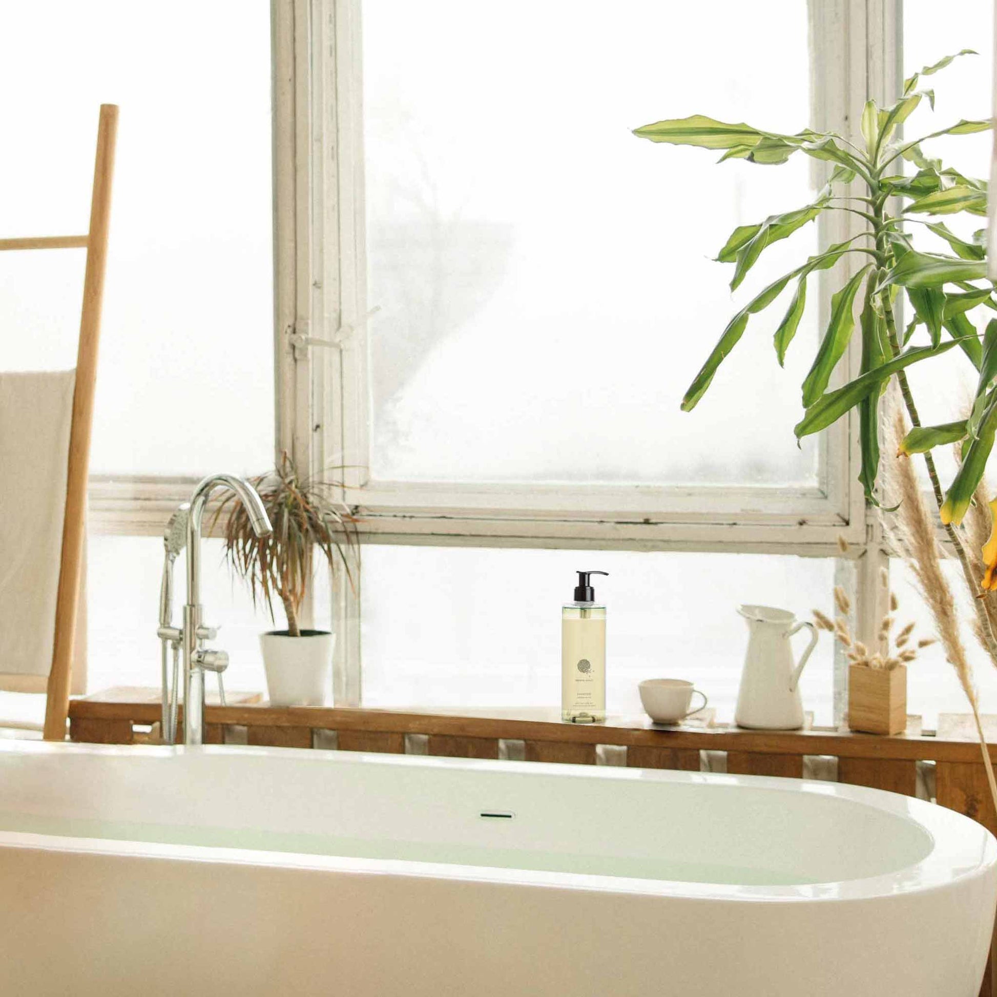 Geneva Guild shampoo 380ml pump dispenser placed on windowsill beside freestanding bathtub in bright, plant-filled hotel bathroom