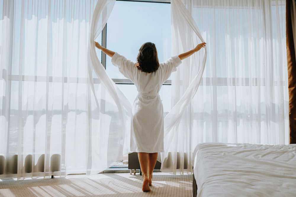 Woman in bathrobe opening blinds, showcasing the comfort and quality of our hotel supplies