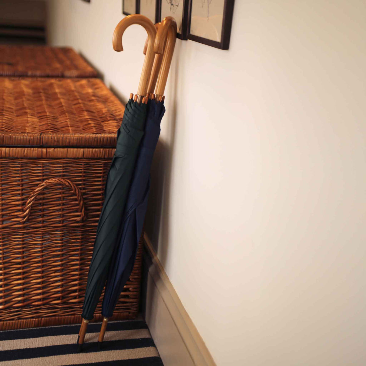 Two closed umbrellas with light wood handles leaning against a white wall next to a woven wicker storage basket in an indoor setting.