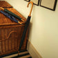 A closed black umbrella with a curved wooden handle leans against a cream-colored wall next to a brown wicker storage basket, with two additional closed umbrellas resting on top of the basket.