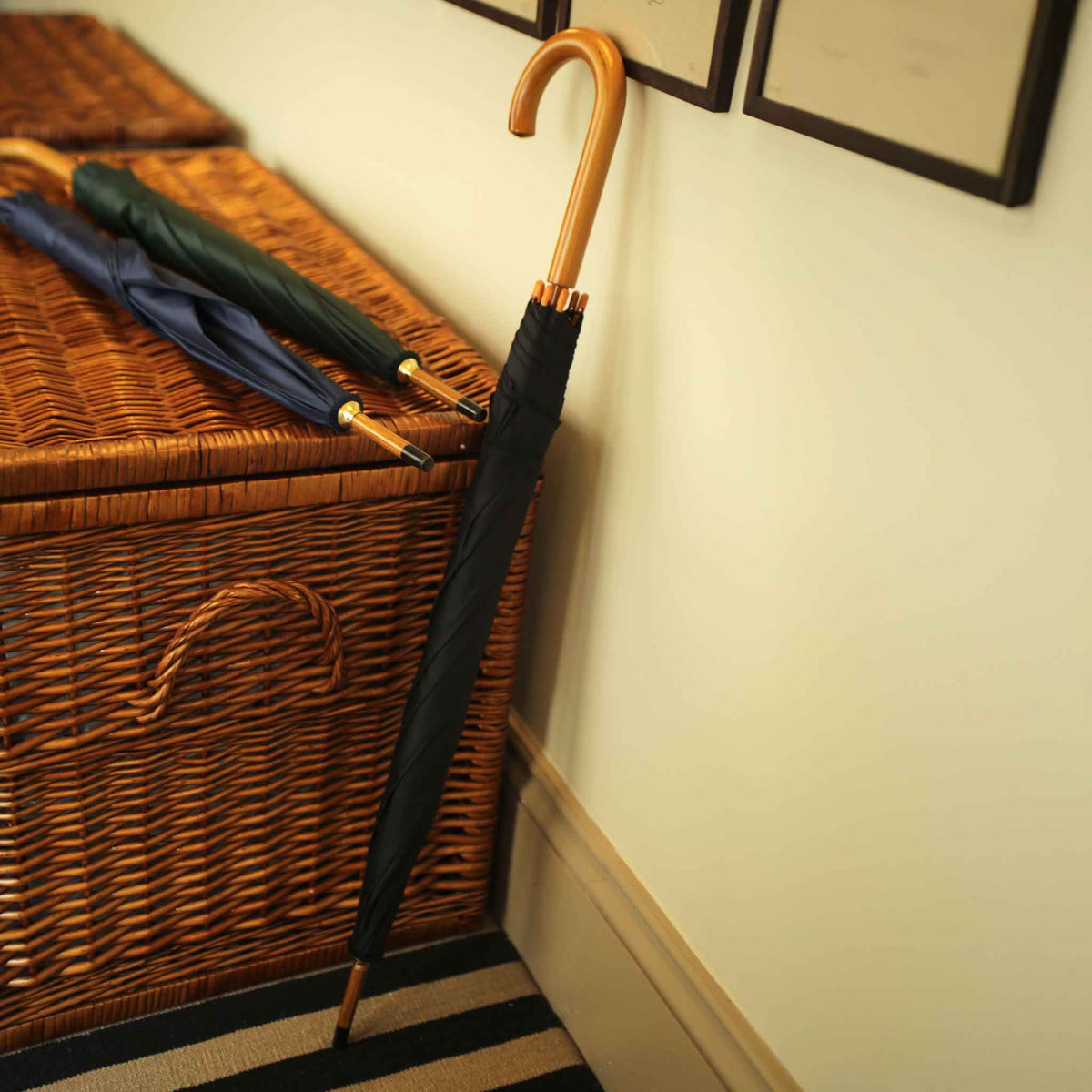 A closed black umbrella with a curved wooden handle leans against a cream-colored wall next to a brown wicker storage basket, with two additional closed umbrellas resting on top of the basket.