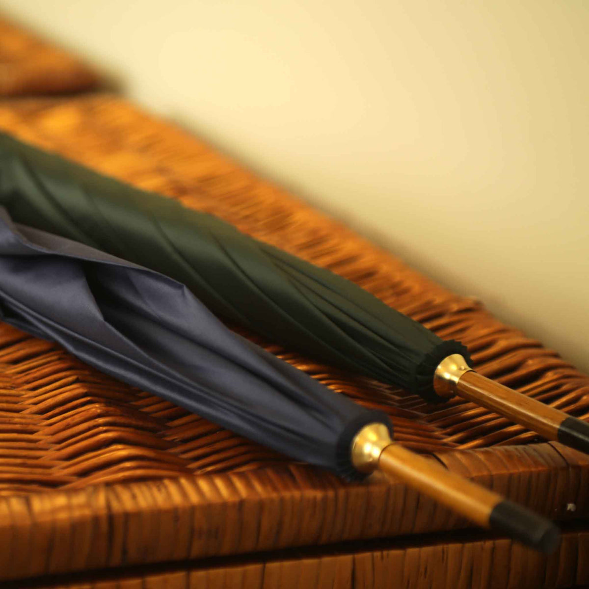 Two closed umbrellas, one dark green and one navy blue, with gold-finished accents, resting diagonally on a brown woven wicker surface.