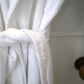 A close-up of a white terry cloth bathrobe tied at the waist, with soft folds and a textured fabric shown against a light neutral background.