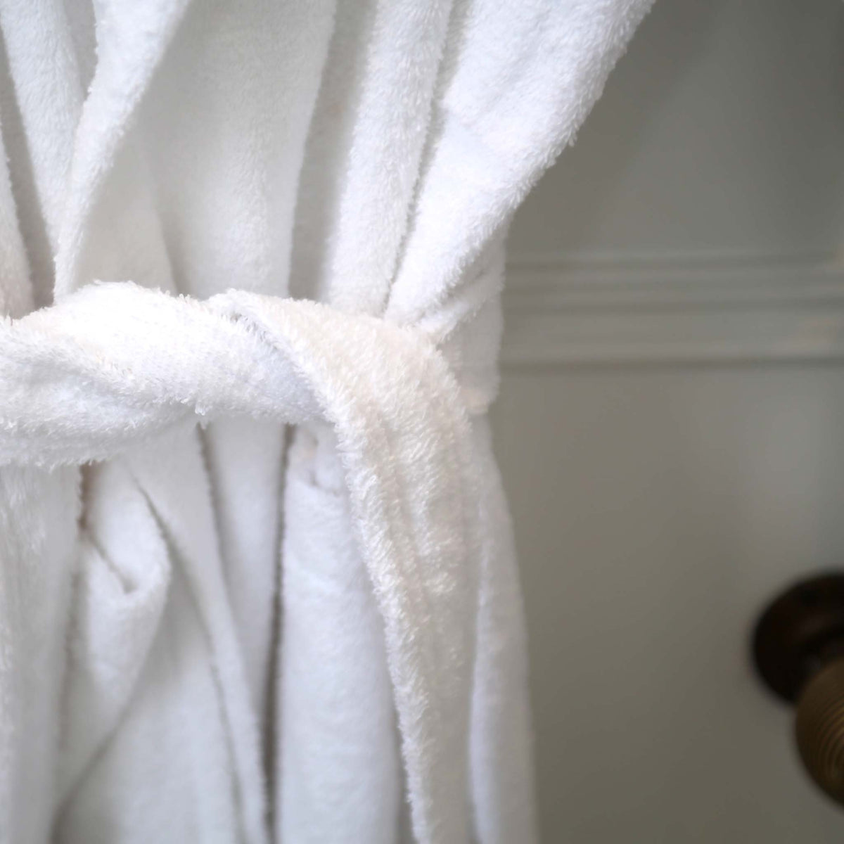 A close-up of a white terry cloth bathrobe tied at the waist, with soft folds and a textured fabric shown against a light neutral background.